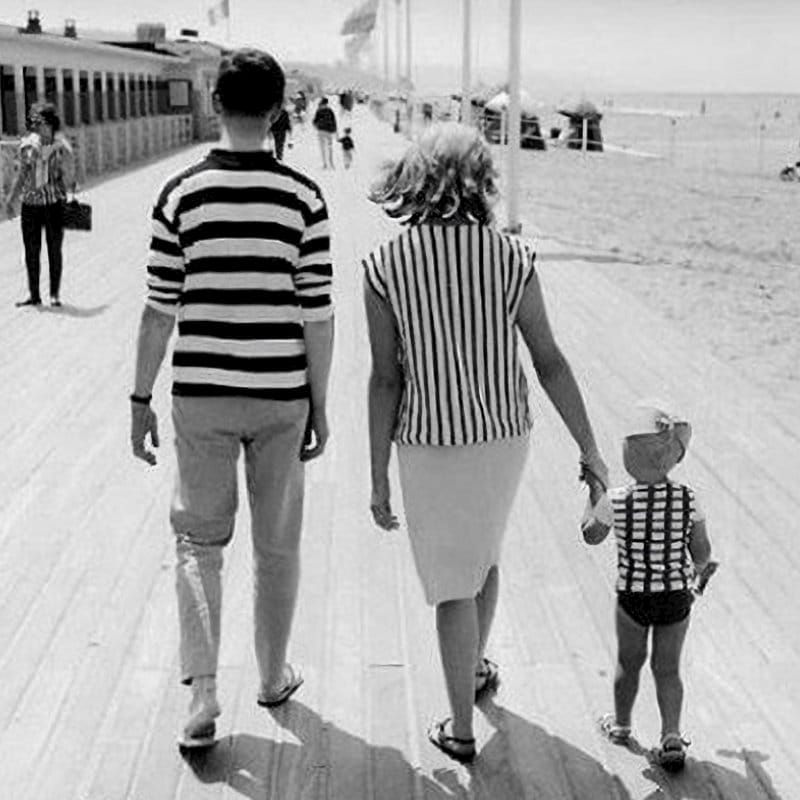 Black-and-white photo of a family walking on a beach boardwalk, with two adults in striped tops and a young child between them wearing a top with combined horizontal and vertical stripes, symbolising inherited genetic traits from both parents.